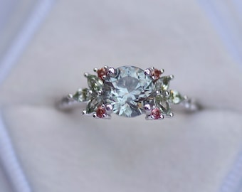 May include: A silver ring with a large, round, light green gemstone surrounded by smaller, green and pink gemstones.