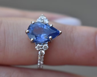 May include: A pear-shaped sapphire engagement ring set in white gold with a halo of smaller diamonds.