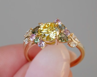 May include: A gold ring featuring a large, round yellow gemstone as the focal point, surrounded by an array of smaller, colorful gemstones in shades of green, pink, and purple. The ring's band is a delicate gold color.