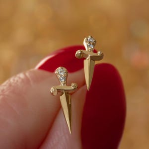 Op de afbeelding: Een paar goudkleurige oorstekers in de vorm van een dolk. Elke oorbel heeft een kleine, sprankelende cluster van heldere edelstenen bij het handvat. De oorbellen worden tegen een vage, warm getinte achtergrond gehouden.