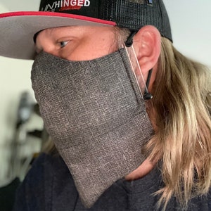 Puede incluir: Una mascarilla gris con un patrón texturizado se usa con una gorra de camionero negra. El sombrero tiene un borde rojo y la palabra "UNHINGED" en letras rojas. Un pin dorado y un pin azul están adheridos al sombrero.