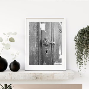 May include: Black and white photograph of an old wooden door with a large ornate metal door handle. The door is weathered and has a keyhole. The photo is framed in a white frame.