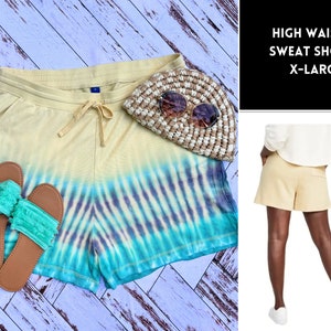 May include: Yellow and blue tie-dye sweat shorts with a high waist and drawstring. The shorts are lying on a white wood surface with a straw hat and two green and brown sandals.