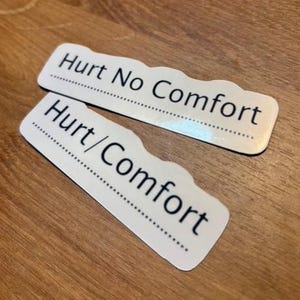May include: Two white stickers with a wavy top edge and the text "Hurt No Comfort" and "Hurt/Comfort" in black. The stickers are on a wooden surface.
