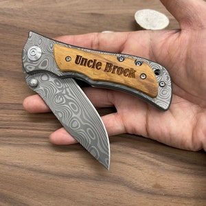 May include: A pocket knife with a wood handle engraved with the words "Uncle Brock". The blade is made of stainless steel with a damascus pattern.