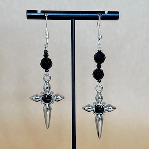 May include: A pair of dangle earrings featuring a silver cross design with a black center stone. The earrings have a silver hook and are accented with black beads. The earrings are displayed on a black stand.