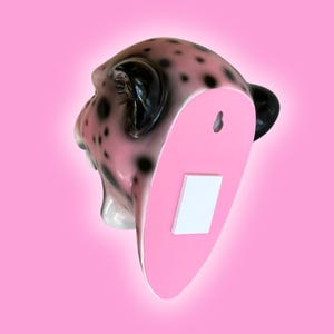 K&ouml;nnte beinhalten: Wanddekoration in Form eines rosa-schwarzen Leopardenkopfes. Der Leopardenkopf hat schwarze Flecken und schwarze Ohren. Die R&uuml;ckseite des Kopfes ist rosa mit einem wei&szlig;en Rechteck und einem Schl&uuml;sselloch zum Aufh&auml;ngen. Der Hintergrund ist einfarbig rosa.