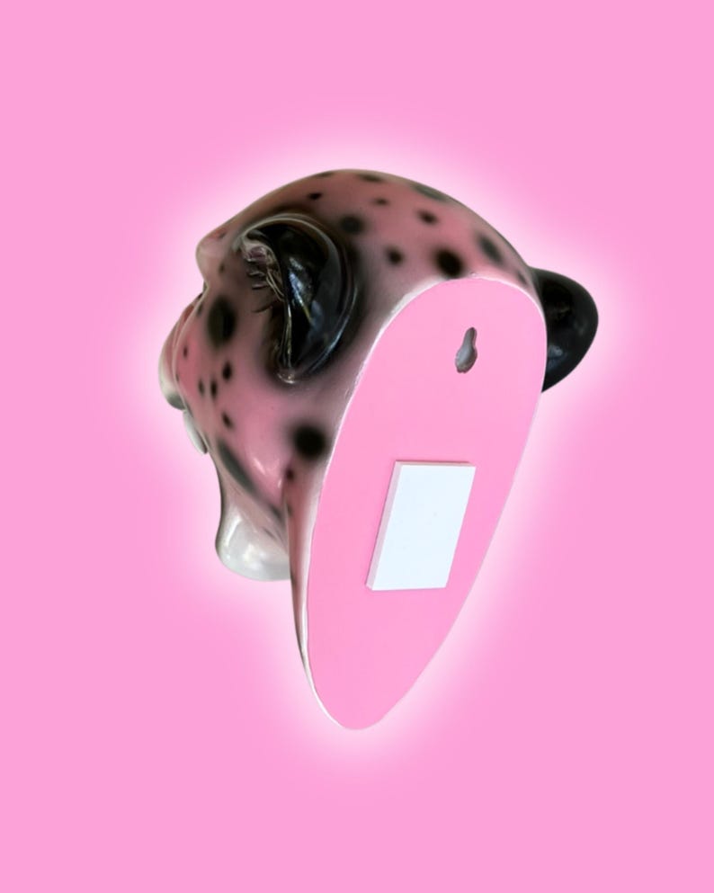 K&ouml;nnte beinhalten: Wanddekoration in Form eines rosa-schwarzen Leopardenkopfes. Der Leopardenkopf hat schwarze Flecken und schwarze Ohren. Die R&uuml;ckseite des Kopfes ist rosa mit einem wei&szlig;en Rechteck und einem Schl&uuml;sselloch zum Aufh&auml;ngen. Der Hintergrund ist einfarbig rosa.