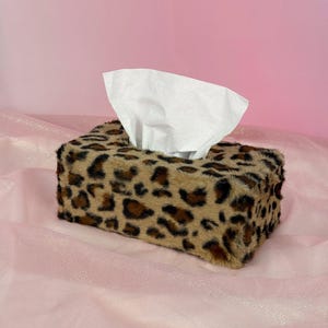 May include: A rectangular tissue box cover featuring a soft, faux fur texture with a brown, black, and tan leopard print pattern. A single white paper tissue is pulled through the top opening of the box, extending upwards from the centre.