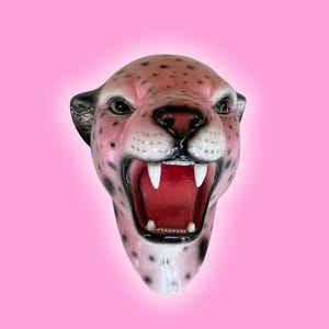 K&ouml;nnte beinhalten: Eine rosa-schwarze Leopardenkopfskulptur mit offenem Maul, wei&szlig;en Z&auml;hnen und rotem Innenraum. Die Skulptur hat schwarze Flecken und gr&uuml;ne Augen, vor einem rosa Hintergrund.