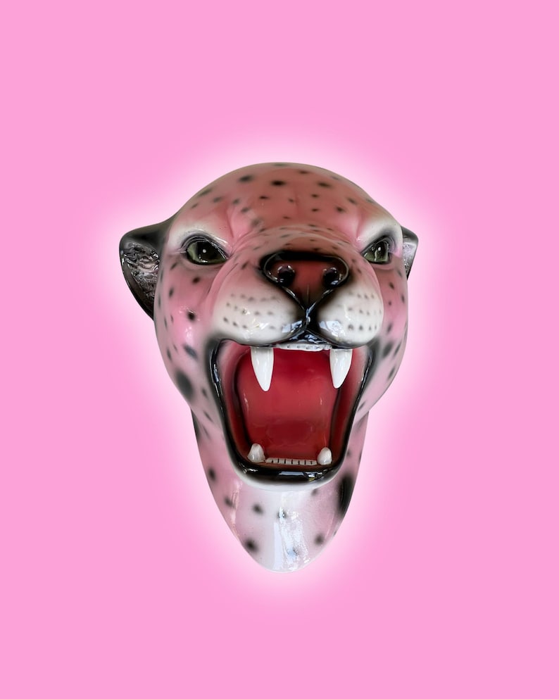 K&ouml;nnte beinhalten: Eine rosa-schwarze Leopardenkopfskulptur mit offenem Maul, wei&szlig;en Z&auml;hnen und rotem Innenraum. Die Skulptur hat schwarze Flecken und gr&uuml;ne Augen, vor einem rosa Hintergrund.