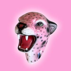 K&ouml;nnte beinhalten: Eine rosa Leopardenkopfskulptur mit schwarzen Flecken, gr&uuml;nen Augen und einem roten, ge&ouml;ffneten Mund, der wei&szlig;e Z&auml;hne zeigt. Die Skulptur hat eine gl&auml;nzende Oberfl&auml;che vor einem rosa Hintergrund.