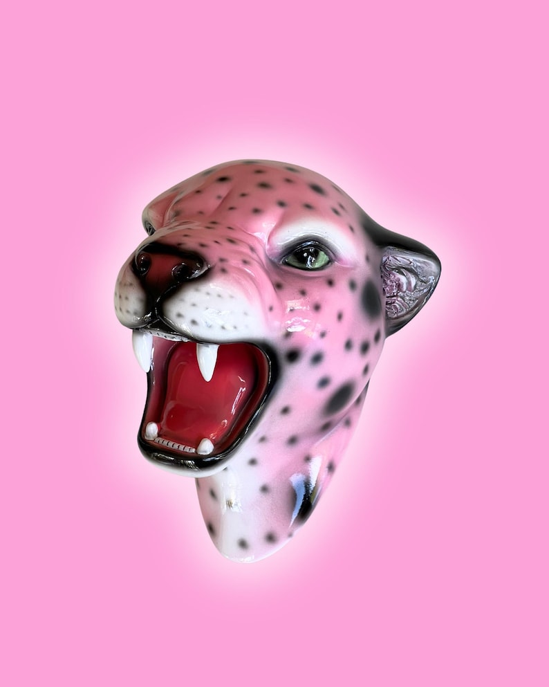 K&ouml;nnte beinhalten: Eine rosa Leopardenkopfskulptur mit schwarzen Flecken, gr&uuml;nen Augen und einem roten, ge&ouml;ffneten Mund, der wei&szlig;e Z&auml;hne zeigt. Die Skulptur hat eine gl&auml;nzende Oberfl&auml;che vor einem rosa Hintergrund.