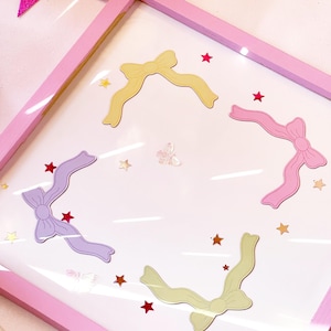 Puede incluir: Una obra de arte enmarcada en rosa con cuatro recortes de cintas en colores pastel: amarillo, rosa, morado y verde claro. Pequeños adornos en forma de estrella roja y dorada están esparcidos alrededor de las cintas. También hay una decoración transparente en forma de mariposa.