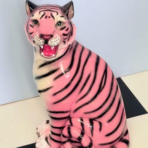 May include: A ceramic tiger figurine featuring a bright pink body with bold black stripes. The tiger is depicted in a seated position with its mouth open, revealing a pink tongue and white teeth. The chest and paws are a creamy white colour.