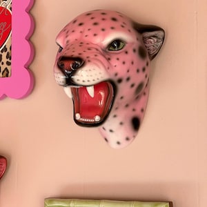 K&ouml;nnte beinhalten: Eine rosa Jaguar-Kopf-Wandskulptur mit schwarzen Punkten, offenem Maul und sichtbaren Z&auml;hnen. Die Skulptur hat gr&uuml;ne Augen und schwarze Akzente an Ohren und Nase. Die Skulptur ist an einer rosa Wand befestigt.