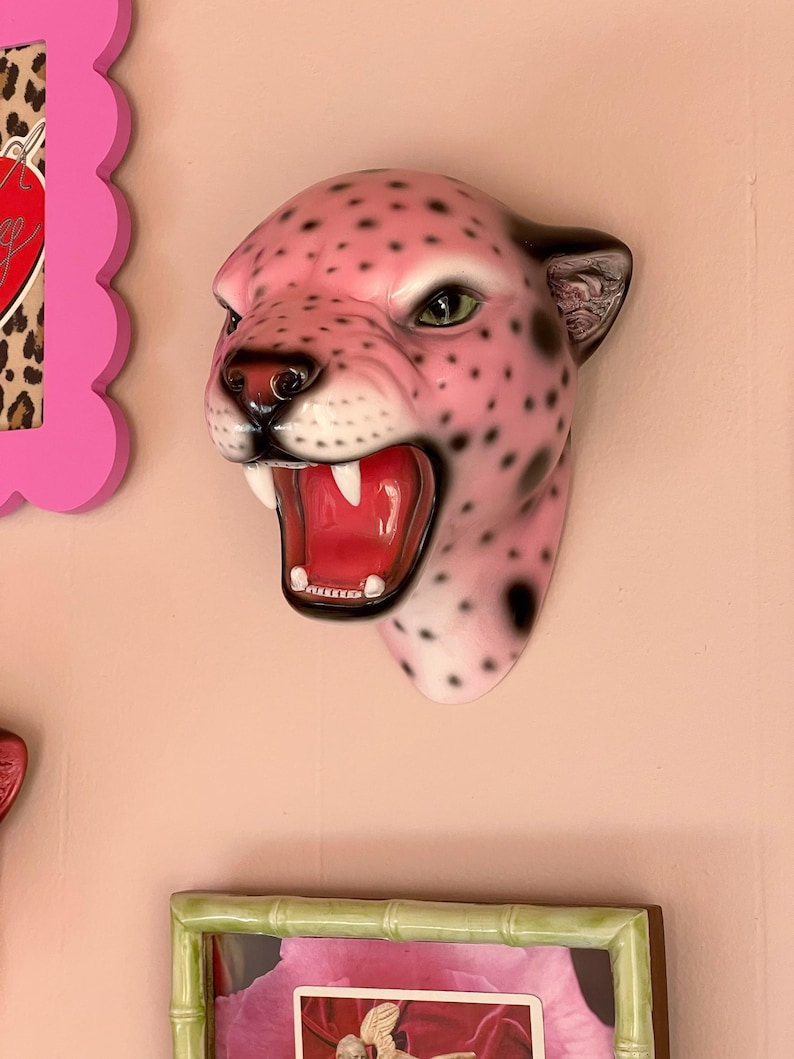 K&ouml;nnte beinhalten: Eine rosa Jaguar-Kopf-Wandskulptur mit schwarzen Punkten, offenem Maul und sichtbaren Z&auml;hnen. Die Skulptur hat gr&uuml;ne Augen und schwarze Akzente an Ohren und Nase. Die Skulptur ist an einer rosa Wand befestigt.