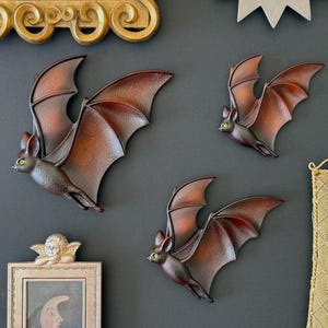 May include: Three wall-mounted bat decorations with textured brown wings and bodies, each featuring small, bright green eyes. The bats are depicted in flight with wings spread wide, showcasing a gradient of dark brown to lighter reddish-brown tones.