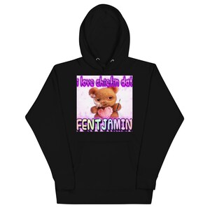 Puede incluir: Sudadera con capucha negra con un gráfico frontal. El gráfico presenta un oso de peluche sosteniendo un corazón y el texto "i love chieflin dat" y "FENTJAMIN" en una fuente colorida. La sudadera tiene una capucha con cordón y un bolsillo delantero.