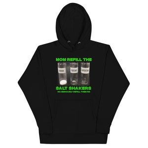 Puede incluir: Sudadera con capucha negra con el texto verde "MOM REFILL THE SALT SHAKERS". Debajo, tres viales transparentes etiquetados "HEROIN", "FENTANYL" y "CARFENTANIL". La frase "NO SERIOUSLY REFILL THEM RN" está en la parte inferior.