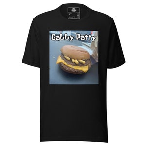 Puede incluir: Camiseta negra con un gráfico de hamburguesa con queso y el texto "Gabby Patty". La hamburguesa tiene dos hamburguesas, queso y mostaza. El texto está en una fuente blanca y juguetona. La camiseta es de material suave.