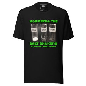 Puede incluir: Camiseta negra con un gráfico humorístico. El diseño presenta tres viales de vidrio etiquetados como "Heroína", "Fentanilo" y "Carfentanilo" con el texto "Mom Refill the Salt Shakers" y "No Seriously Refill Them RN" en verde.