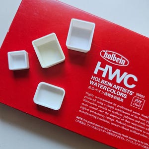 Könnte beinhalten: Eine rote Schachtel mit Holbein Artists' HWC Aquarellfarben mit dem Text "HOLBEIN ARTISTS' W401 WATERCOLORS". Mehrere kleine, weiße, leere Aquarellfarben-Näpfe sind auf der Schachtel angeordnet. Die Schachtel enthält auch japanischen Text.