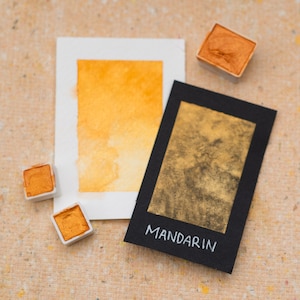May include: A watercolor paint pan with a sample of the color Mandarin. The paint is a bright orange color with a metallic sheen. The pan is labeled with the word "Mandarin" in white lettering.