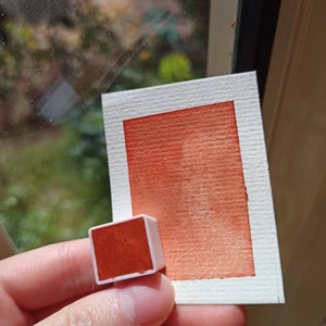 May include: A small, square pan of orange watercolor paint. The pan is in a white plastic case. A sample of the paint is shown on a white card.