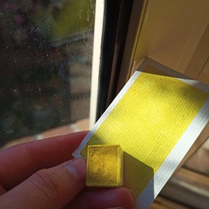 May include: A small, square, yellow watercolor paint pan. The pan is sitting on a white paper with a yellow watercolor wash.
