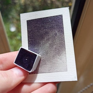 May include: A small, square watercolor pan with a dark purple pigment. The pan is sitting on a white card with a purple watercolor wash.