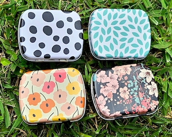 May include: Four small, rectangular metal tins with different patterns. The tins have a black and white polka dot pattern, a teal leaf pattern, a floral pattern with pink and white flowers on a black background, and a yellow and orange flower pattern on a white background.