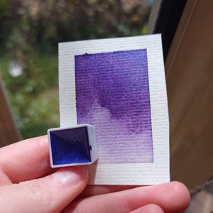May include: A small, square watercolor pan with a purple pigment. The pan is sitting on a white card with a purple watercolor wash.