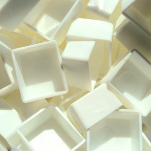 May include: A close-up of a pile of white plastic watercolour paint pans. The pans are square and have a small well in the centre.