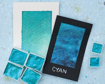 May include: A set of cyan watercolor paints in individual pans. The pans are arranged in a square shape. A watercolor wash of cyan is painted on a white card with the word "CYAN" written in white on a black card.