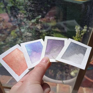 May include: Four watercolor paint swatches on white card stock. The swatches are arranged in a fan shape. The colors are a shimmery orange, a shimmery blue, a shimmery purple, and a gray.