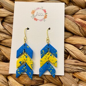 Puede incluir: Un par de pendientes de chevron azul y amarillo con detalles dorados. Los pendientes est&aacute;n hechos de arcilla polim&eacute;rica y tienen un acabado moteado.