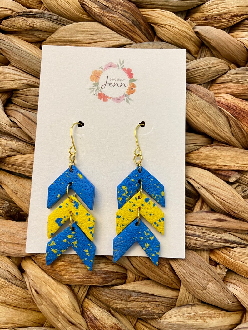 Puede incluir: Un par de pendientes de chevron azul y amarillo con detalles dorados. Los pendientes est&aacute;n hechos de arcilla polim&eacute;rica y tienen un acabado moteado.