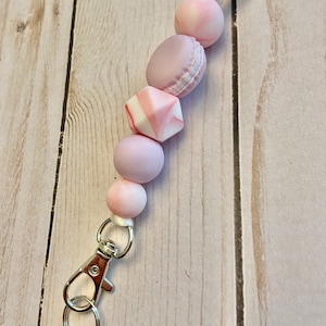 May include: A pink and white beaded keychain with a silver lobster clasp. The beads are in the shape of macarons and a square.