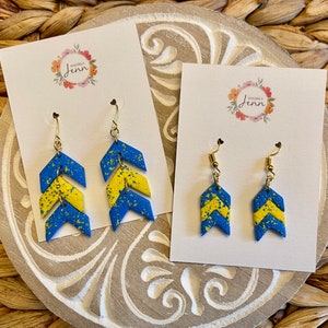 Puede incluir: Dos pares de pendientes de chevron azul y amarillo con ganchos dorados. Los pendientes se muestran en tarjetas blancas con el texto "Sincerely Jenn" en cada tarjeta.