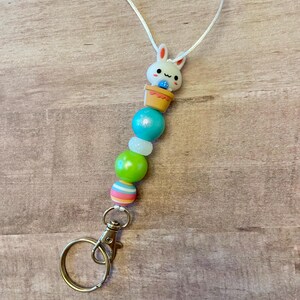 May include: A white bunny charm with a blue egg in a yellow pot, attached to a white cord with a silver key chain and colorful beads. The beads are green, white, and striped.