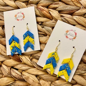 Puede incluir: Un par de pendientes de chevron azul y amarillo con ganchos dorados. Los pendientes est&aacute;n hechos de madera y tienen un acabado moteado. Los pendientes se muestran en una tarjeta blanca con el texto "Sincerely Jenn" en un dise&ntilde;o floral.
