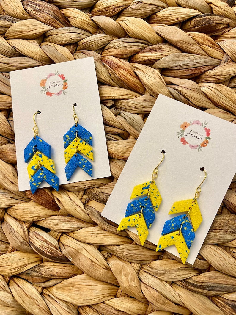 Puede incluir: Un par de pendientes de chevron azul y amarillo con ganchos dorados. Los pendientes est&aacute;n hechos de madera y tienen un acabado moteado. Los pendientes se muestran en una tarjeta blanca con el texto "Sincerely Jenn" en un dise&ntilde;o floral.