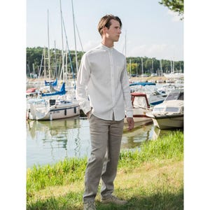 May include: A long-sleeved white button-down shirt with a mandarin collar and a single chest pocket, paired with straight-leg light brown trousers. The shirt features a full-length front button placket and is worn tucked into the trousers.
