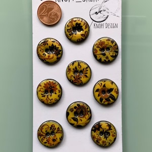 May include: Nine yellow, floral-patterned buttons with brown and orange detailing. The buttons are round with two holes and a glossy finish. A 2 Euro cent coin is visible. The card reads "KNOPF DESIGN".