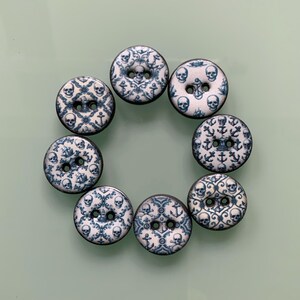 Könnte beinhalten: Ein Set aus sieben runden Knöpfen, die in einem Kreis angeordnet sind. Jeder Knopf hat eine weiße Basis mit einem wiederkehrenden blauen Muster aus Totenköpfen, Ankern und komplexen Schnörkeln. Jeder Knopf hat zwei mittige Löcher.