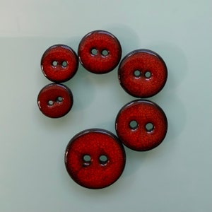 Könnte beinhalten: Sechs rote Keramikknöpfe mit jeweils zwei Löchern. Die Knöpfe sind in einem Halbkreis angeordnet.