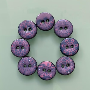 May include: A set of seven circular buttons arranged in a circle. Each button features a two-hole design and a dark purple surface decorated with a repeating intricate pink floral pattern. The buttons have a smooth, glossy finish and uniform shape.