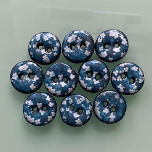 Peut inclure: Dix boutons ronds avec un fond bleu foncé et un motif floral blanc. Chaque bouton comporte deux trous pour la couture. Les boutons sont disposés en grille. Les boutons mesurent environ 2 cm de diamètre.