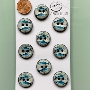 Könnte beinhalten: Zehn runde Knöpfe mit Fischmuster. Jeder Knopf hat eine blau-weiße Illustration von Fischen, die im Kreis schwimmen. Die Knöpfe befinden sich auf einer weißen Karte mit dem Text "KNOPF DESIGN". Eine 2-Cent-Euro-Münze befindet sich in der oberen linken Ecke.